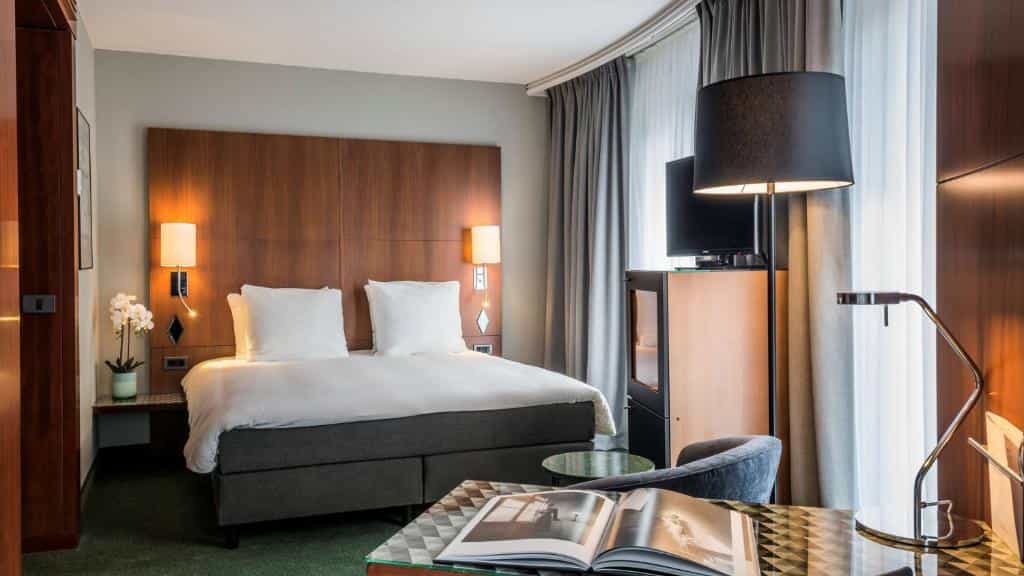 Hotel bedroom with large bed, wooden headboard, floor lamp, and open book on desk.