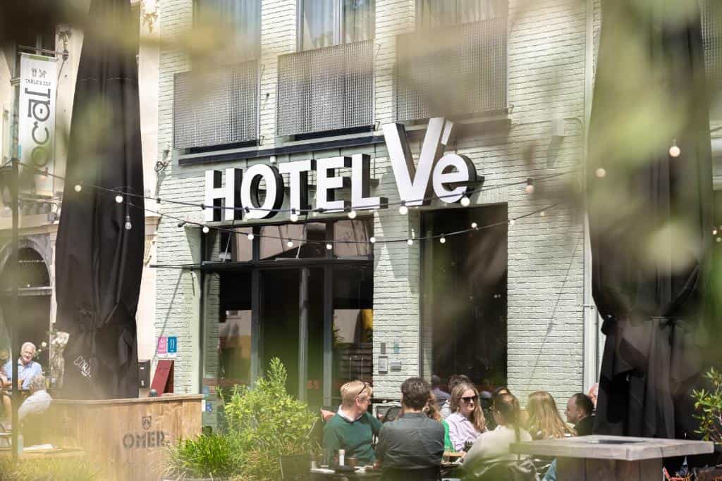 Outdoor dining area in front of Hotel Vé with patrons seated near a light green brick building.