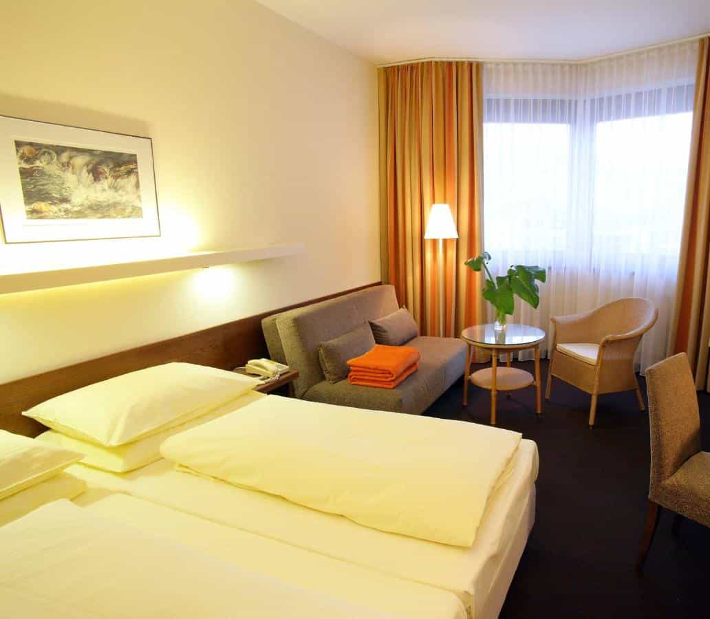 Hotel room with twin beds, sofa, wicker chair, and bright window with orange curtains in Hotel Sonne Lienz.