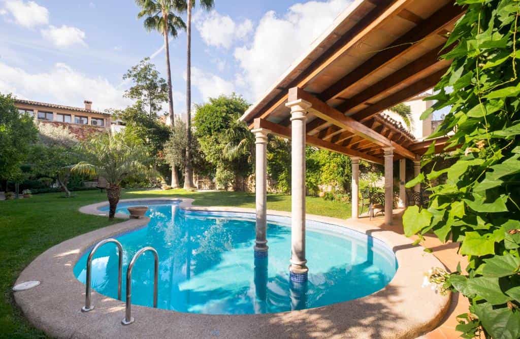 Swimming pool with curved edge, covered pergola, and lush green garden with palm trees.