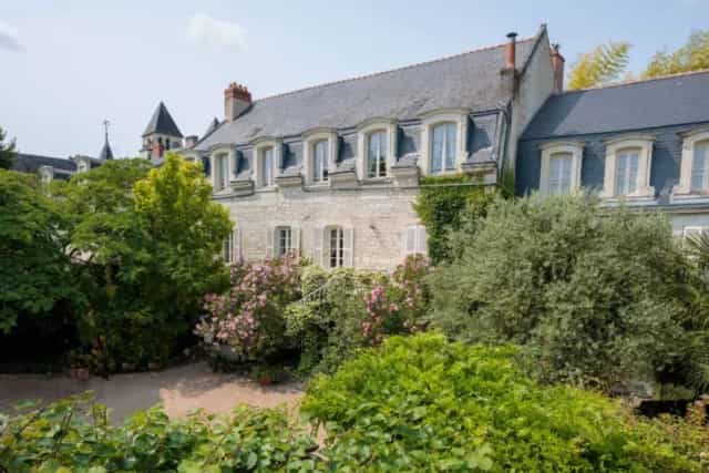 Stone building facade with a slate roof, surrounded by lush green trees and flowering bushes.