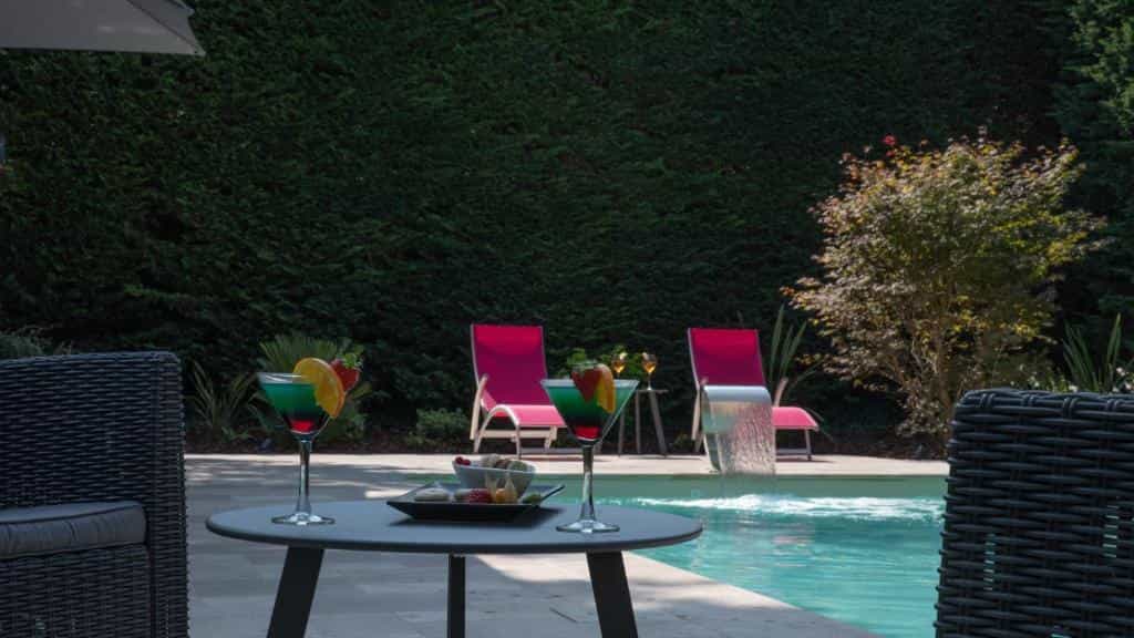 Cocktails and snacks on a table by a swimming pool with lounge chairs and a garden hedge.