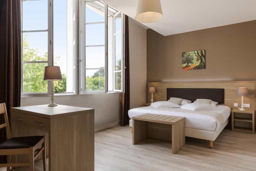 Hotel room with a double bed, wooden desk, and large open windows overlooking trees.