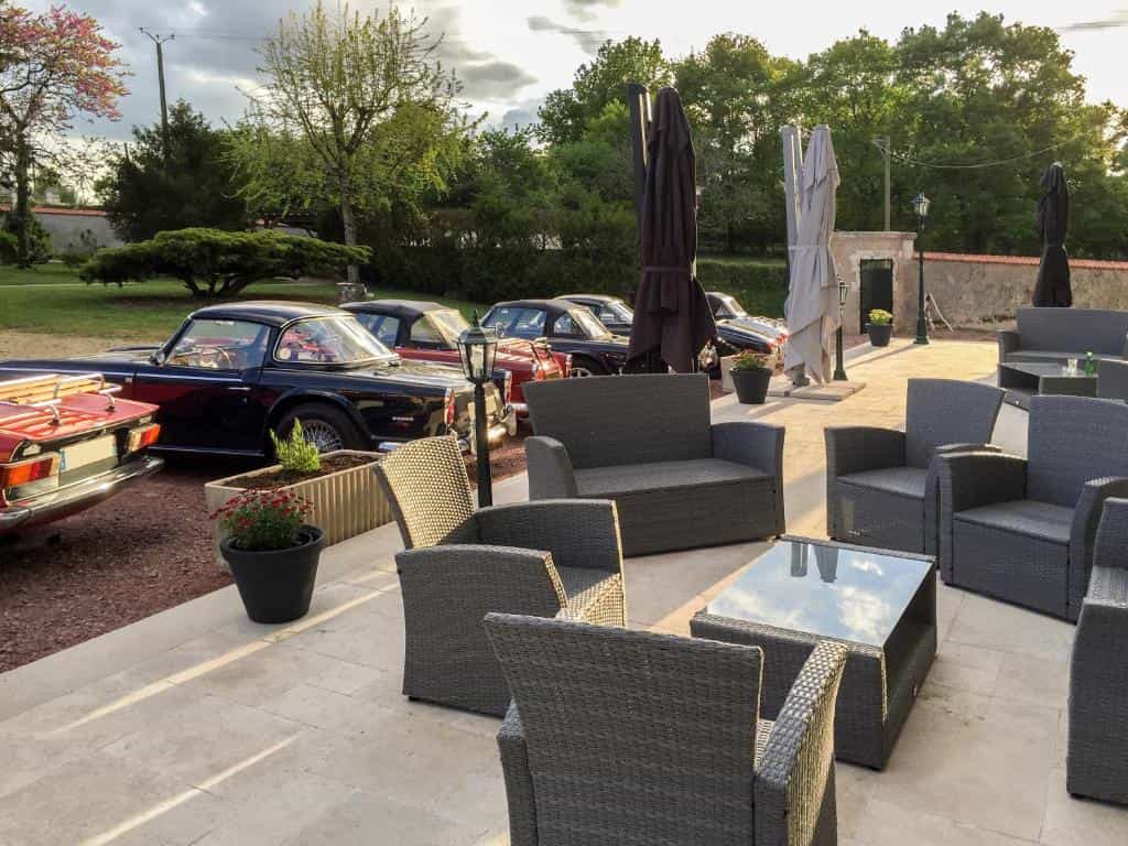 Vintage convertible cars parked on a gravel lot next to an outdoor patio with wicker furniture.