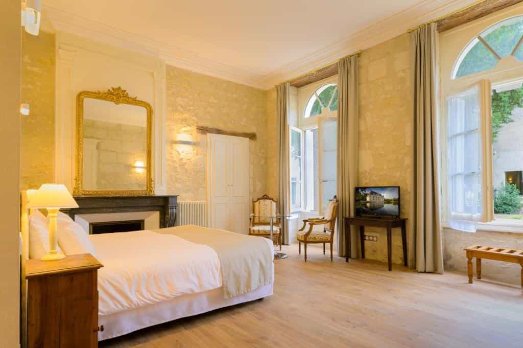 Hotel bedroom with stone walls, fireplace, ornate gold mirror, and large windows overlooking a courtyard.