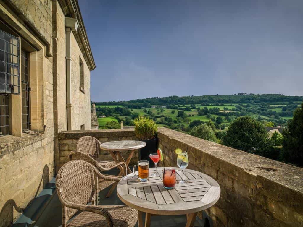 Outdoor cafe tables with drinks on a stone terrace overlooking a green countryside landscape.