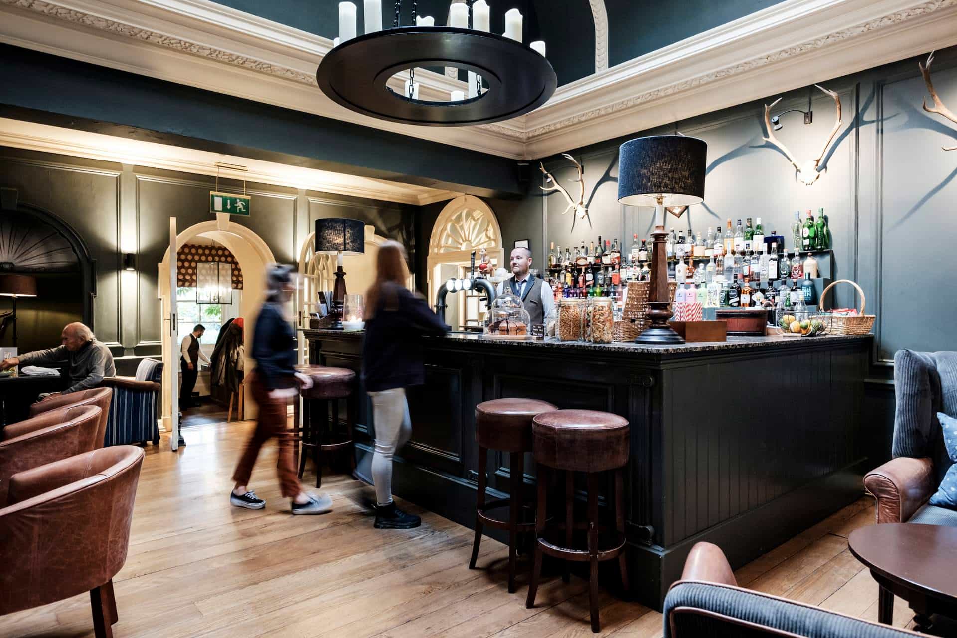 People at a dark wood bar in a traditional pub with leather stools and antler wall decor.