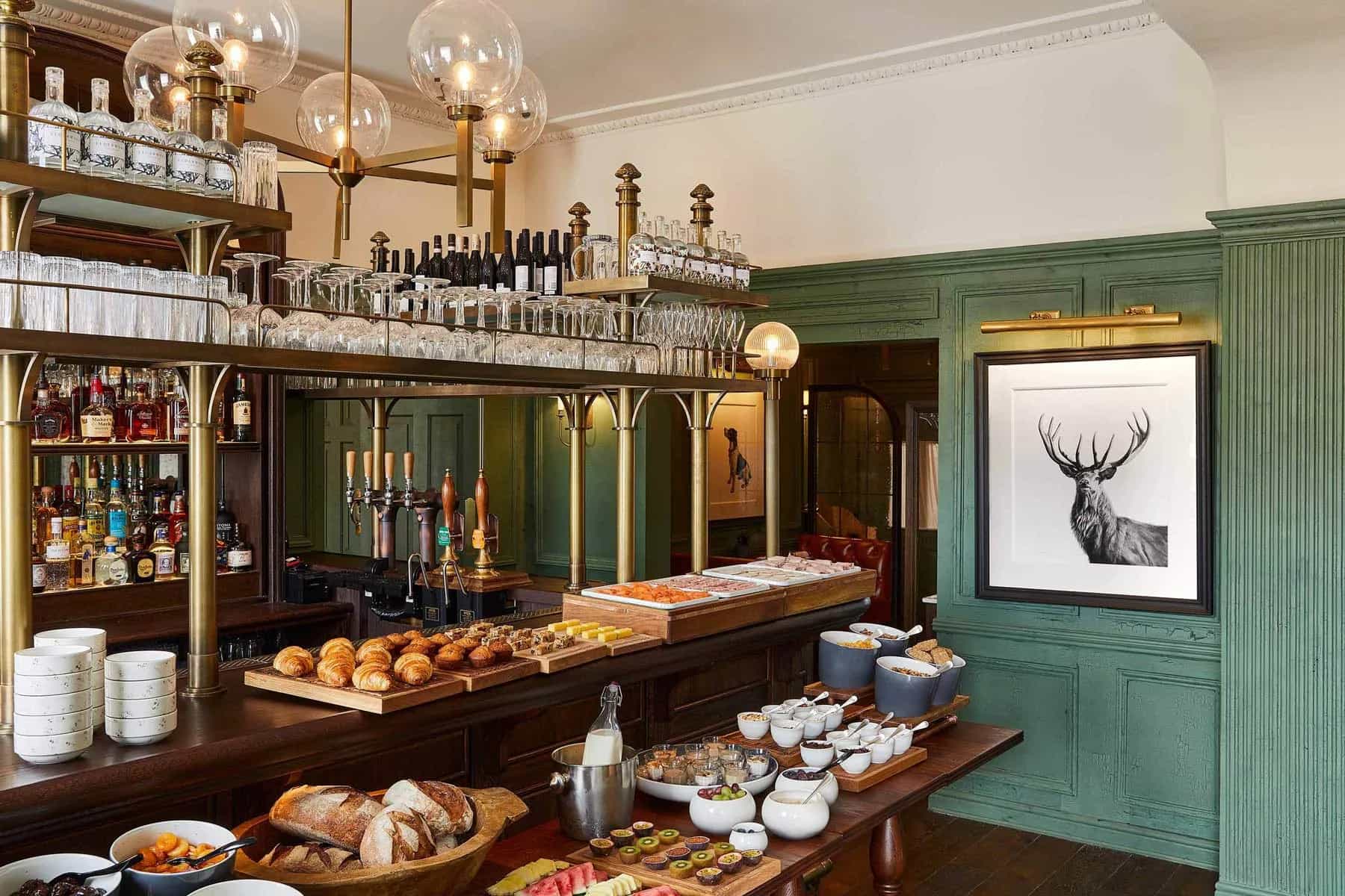Breakfast buffet spread on a wooden bar counter in a pub with green paneled walls and a stag print.