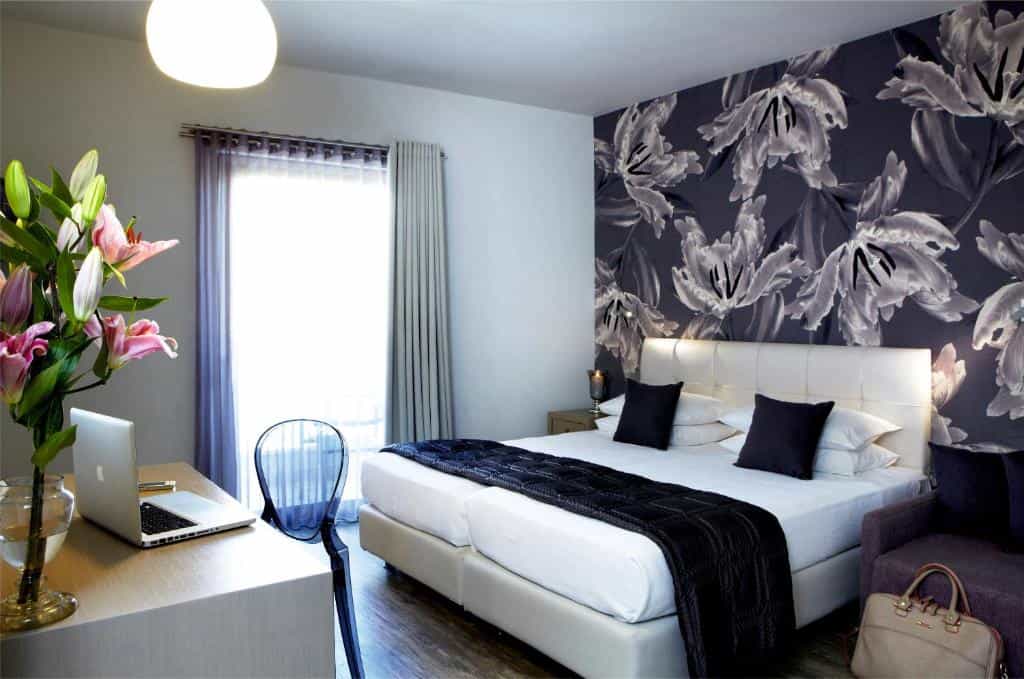 Hotel room with a double bed, floral accent wall, desk with a laptop, and a vase of lilies.