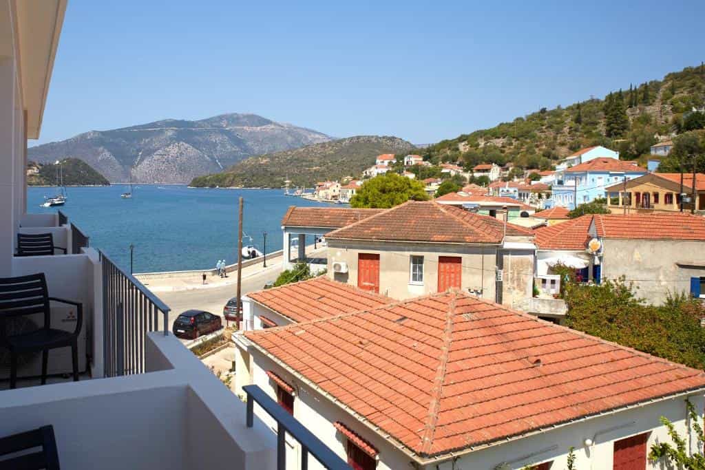 Coastal village with terracotta roofs overlooking a bay with sailboats and mountains.