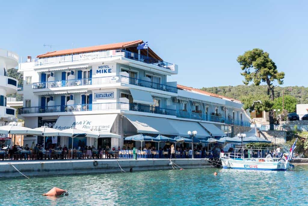 Hotel Mike and restaurant with outdoor seating by the harbor in Greece with a boat docked in the water.