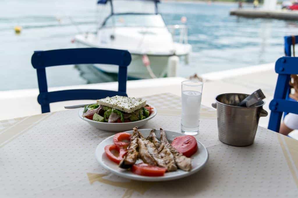 Greek salad and grilled fish on a table at a waterfront restaurant with a boat in the background.