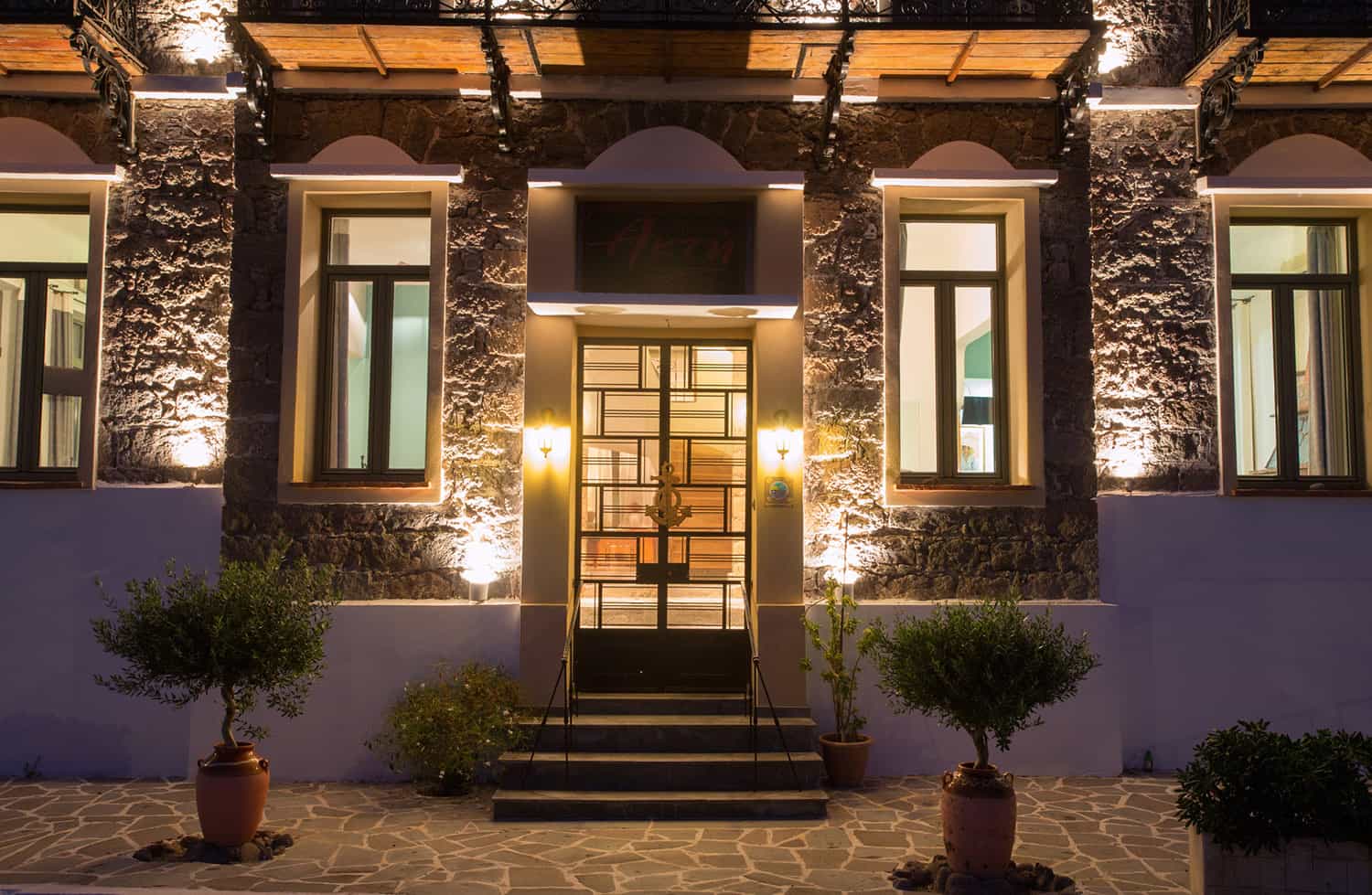 Illuminated stone building entrance with potted olive trees at night.