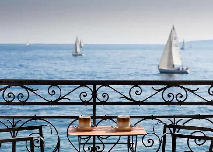 Two coffee cups on a small table on a balcony overlooking sailboats on the sea.