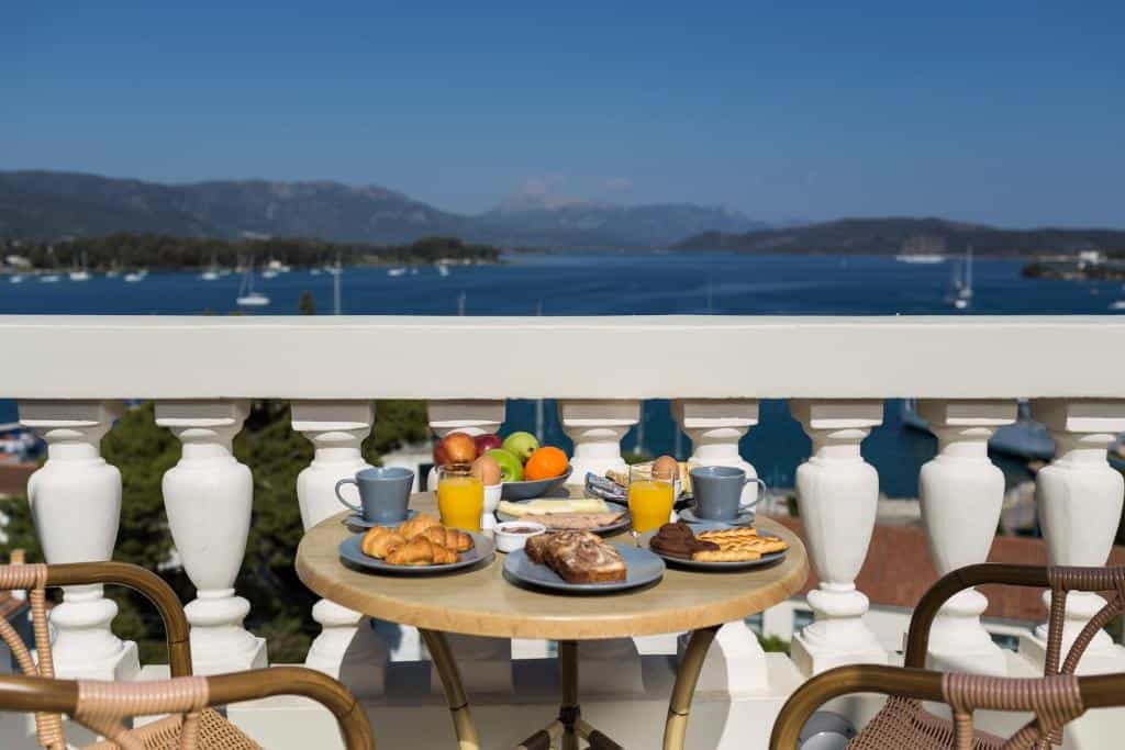 Breakfast table with fruit, juice, and pastries on a balcony overlooking a bay with sailboats and mountains.