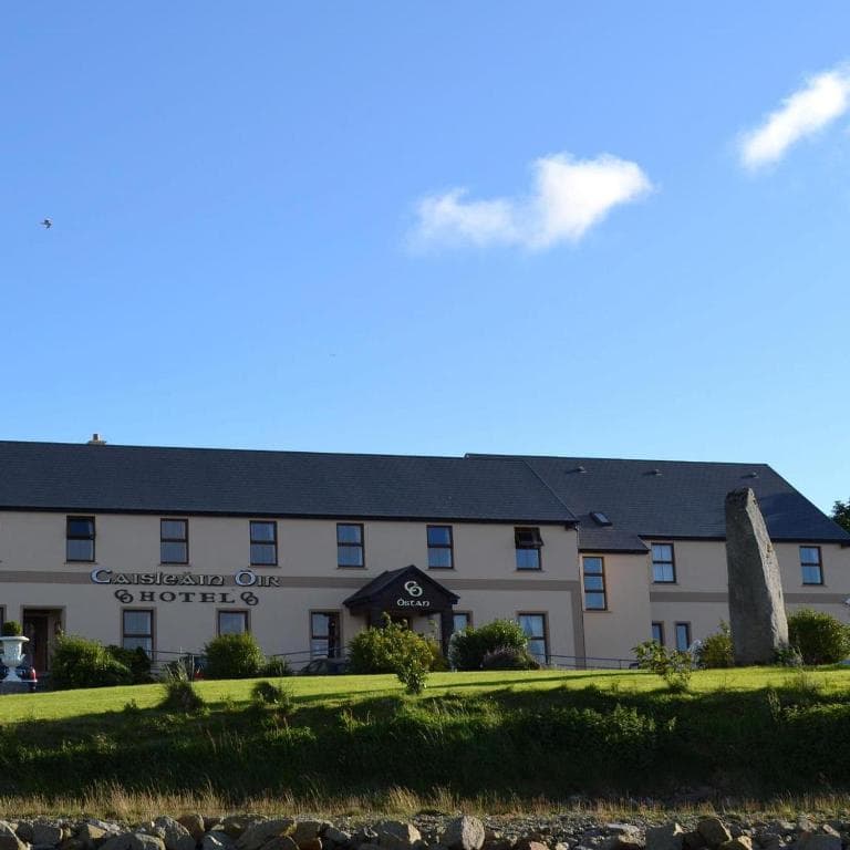 Caisleain Oir Hotel building with sign and large standing stone on grassy lawn under blue sky.