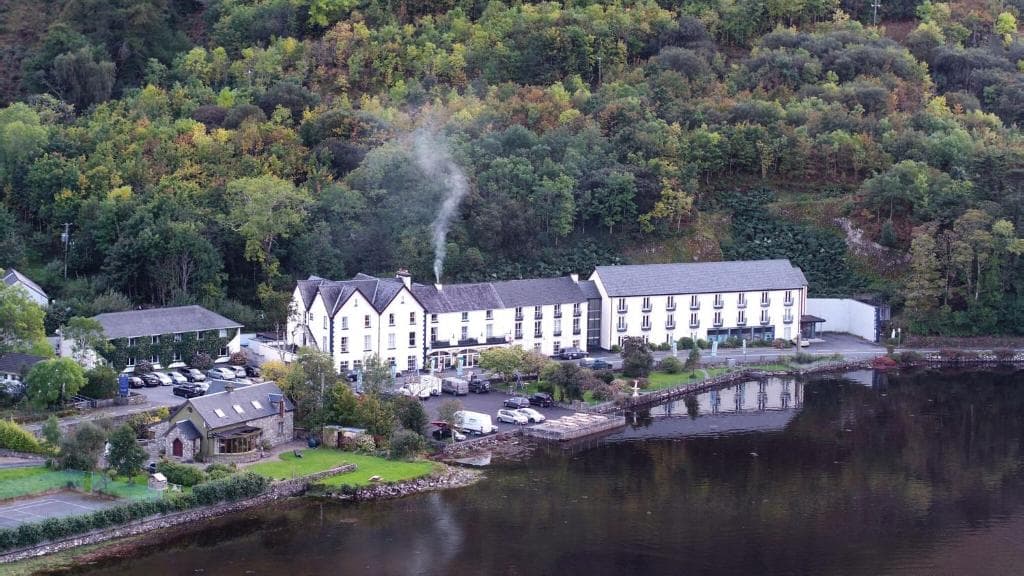 White hotel building by dark water with smoke rising, backed by dense green hillside.