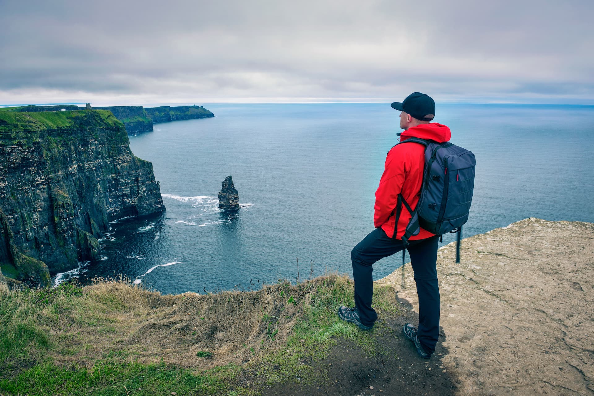 Hiker viewing Cliffs of Moher sea stack and dramatic coastal cliffs under cloudy sky.