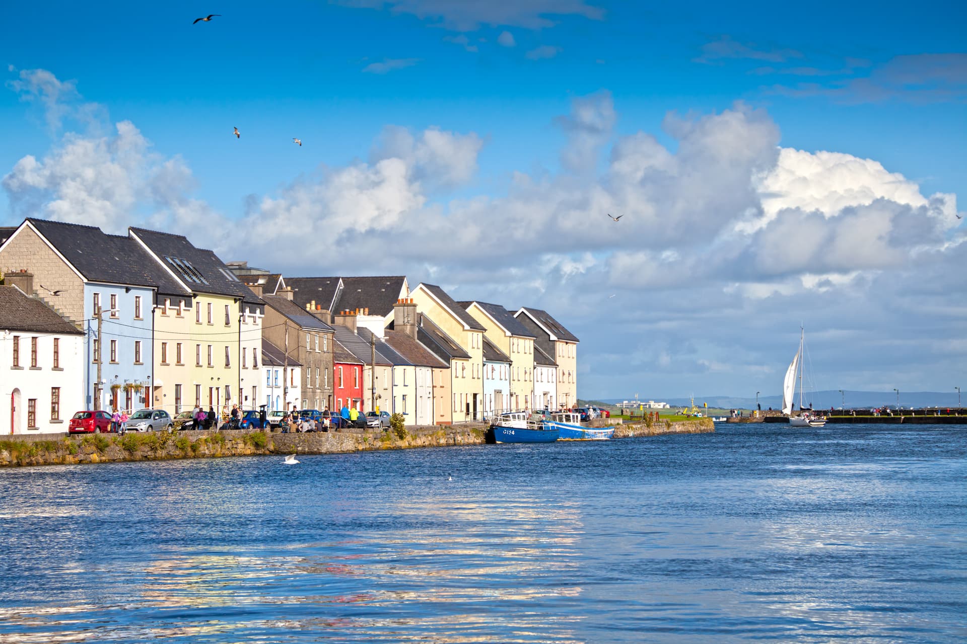 Colorful waterfront buildings along the water in Galway City with a sailboat on the water.
