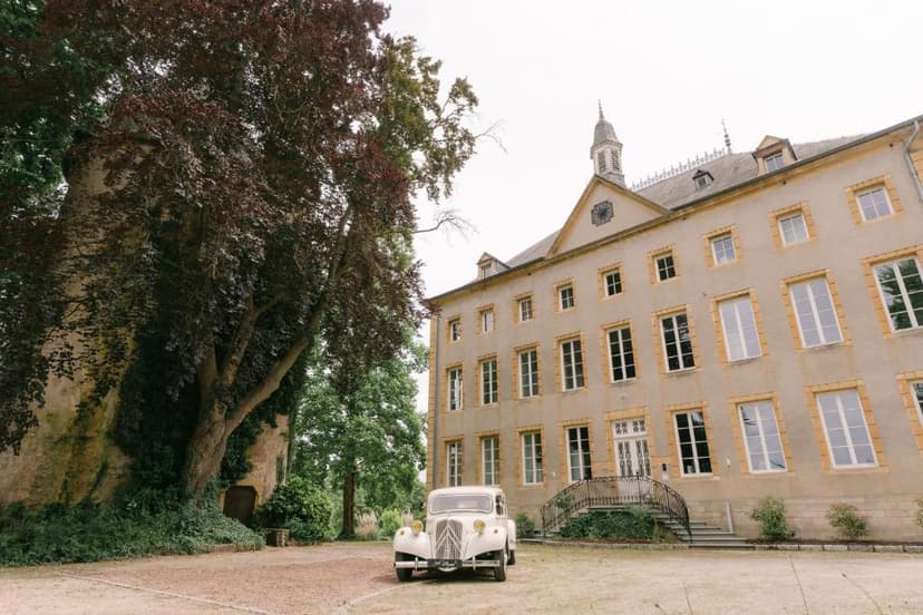 Vintage white car parked before large historic building with large tree in Remich.