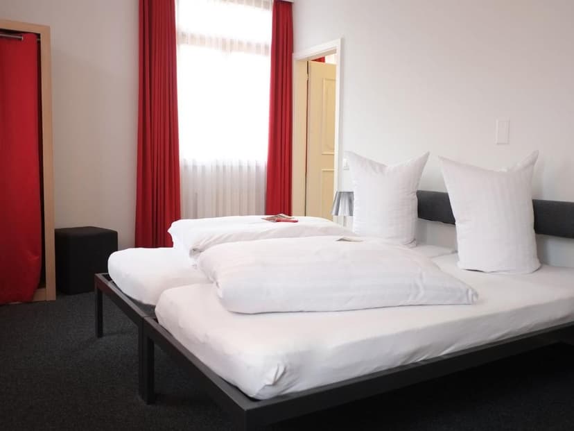 Twin beds with white linens in a bright room with red curtains at Hotel Brauerei Frohsinn Arbon.