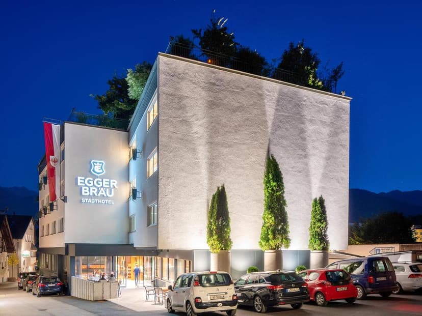 Stadthotel Eggerbräu illuminated at night with parked cars and mountains in background, Imst