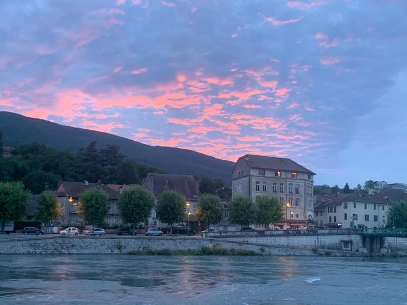 Riverfront buildings in Seyssel at sunset with pink clouds over dark mountains.