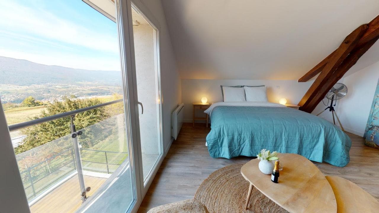 Attic bedroom with blue quilt, wooden beams, and balcony view of valley near Seyssel.