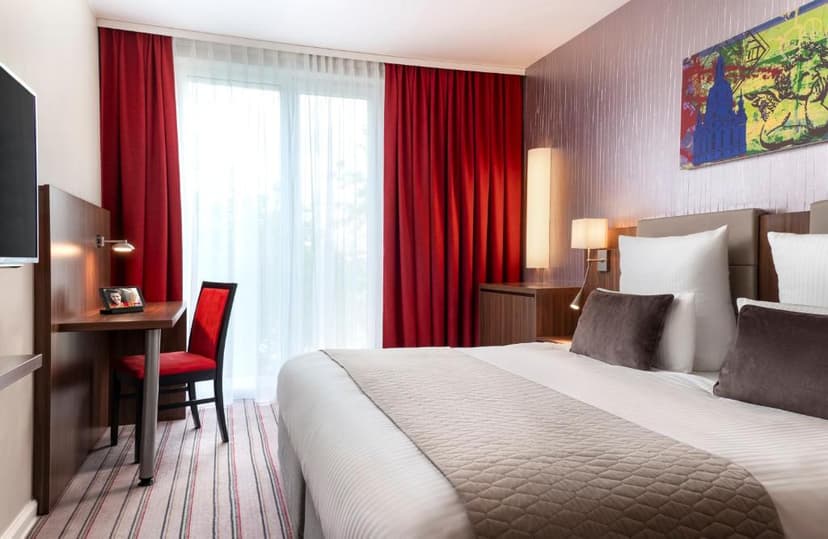 Hotel room with white bedding, red curtains, and modern artwork featuring a building silhouette.