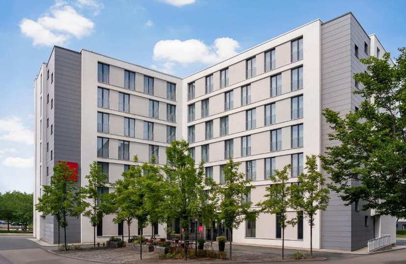 Modern hotel building exterior with gray and white facade, green trees, and blue sky.