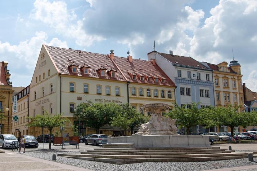 Hotel Česká Koruna and fountain on cobblestone square with parked cars under cloudy sky.