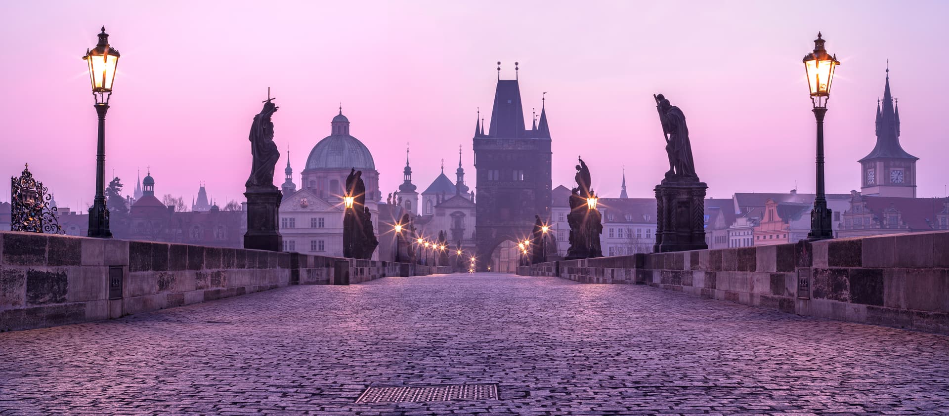 Cobblestone Charles Bridge statues and glowing lamps at purple sunrise in Prague.
