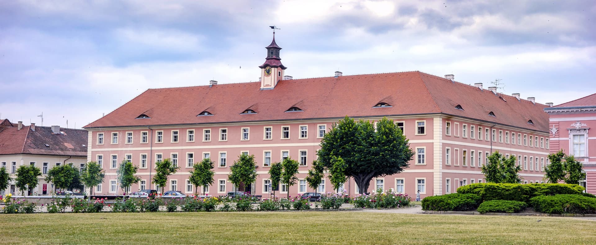 Large pink historic building with clock tower and red tile roof overlooking a grassy park in Terezín.