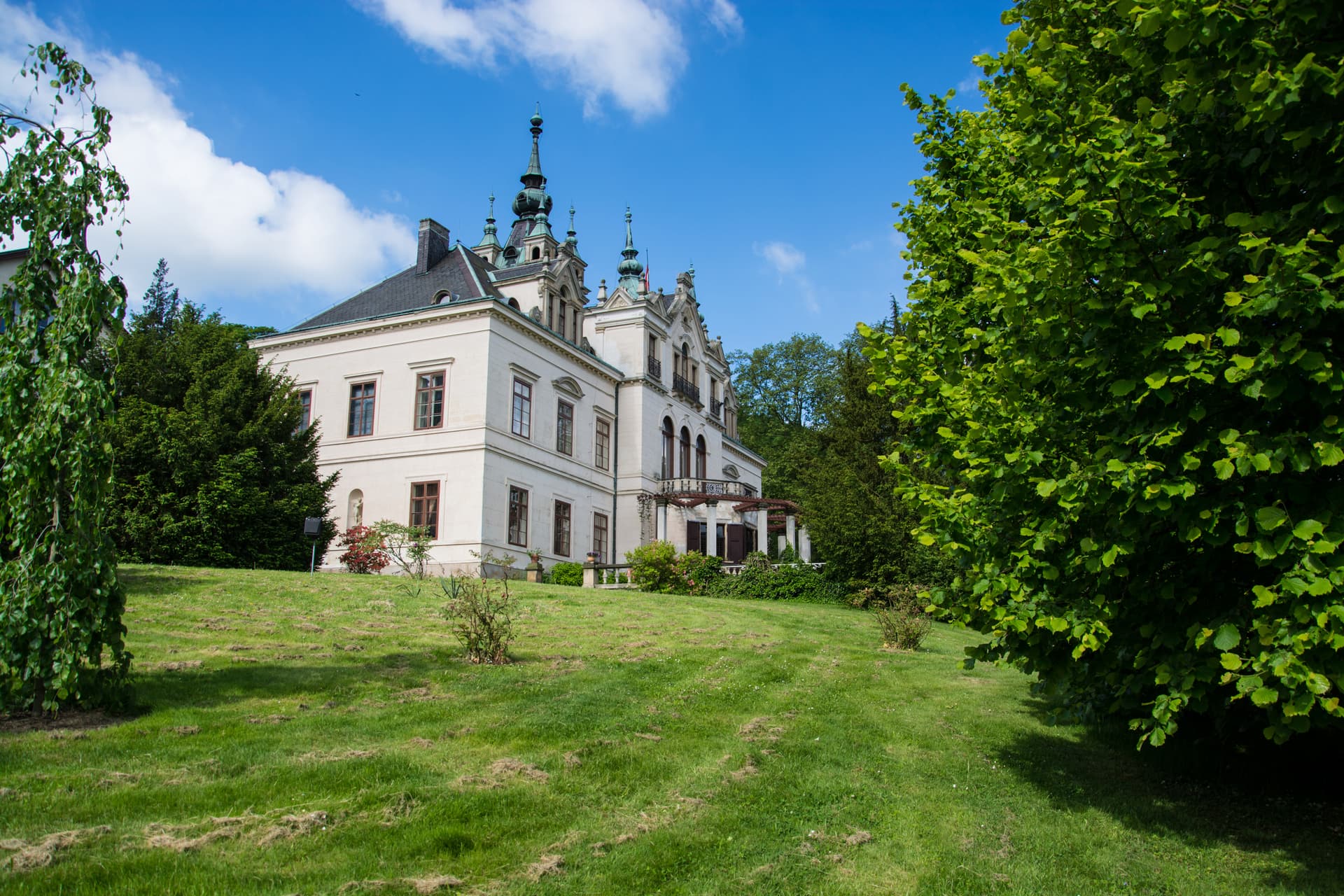Ornate white manor house with green spires on a grassy hill under a blue sky in Velké Březno.