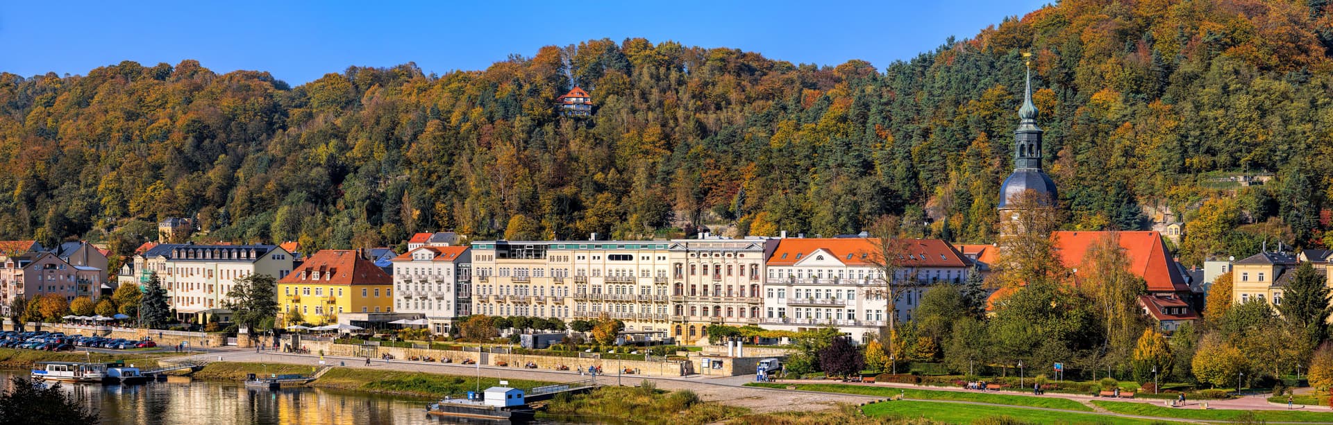 Bad Schandau town with historic buildings, river, and autumn-colored forest hills under blue sky.