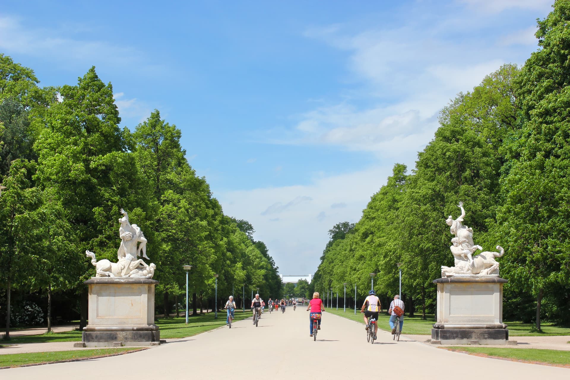 Cyclists riding down wide promenade lined with lush green trees and classical statues, Grosser Garten