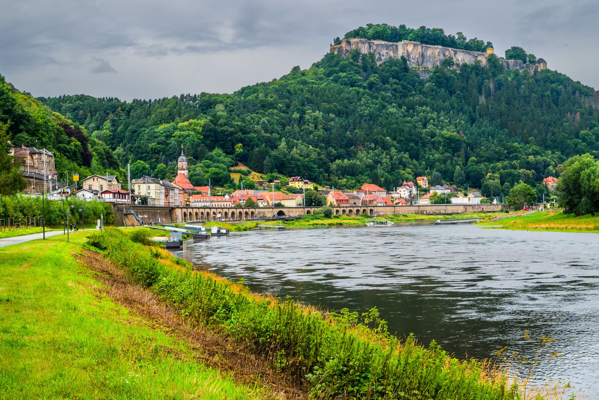 River town with buildings along the water, forest, and Koenigstein Fortress on the cliff.