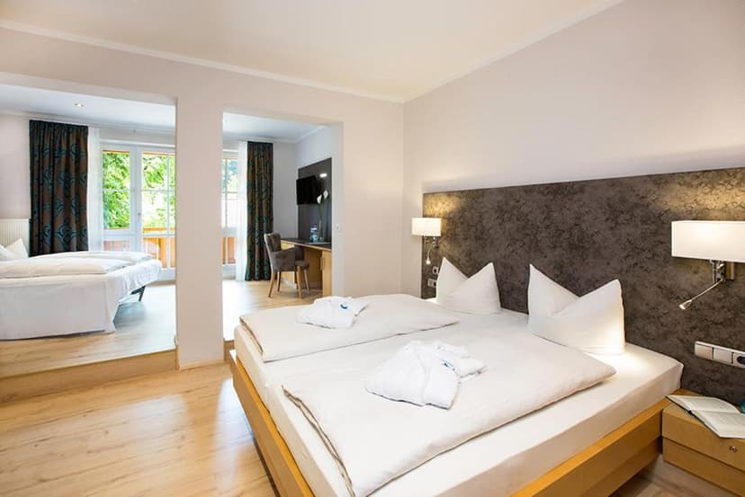 Hotel room suite with two beds, light wood floors, and view of greenery through balcony doors.