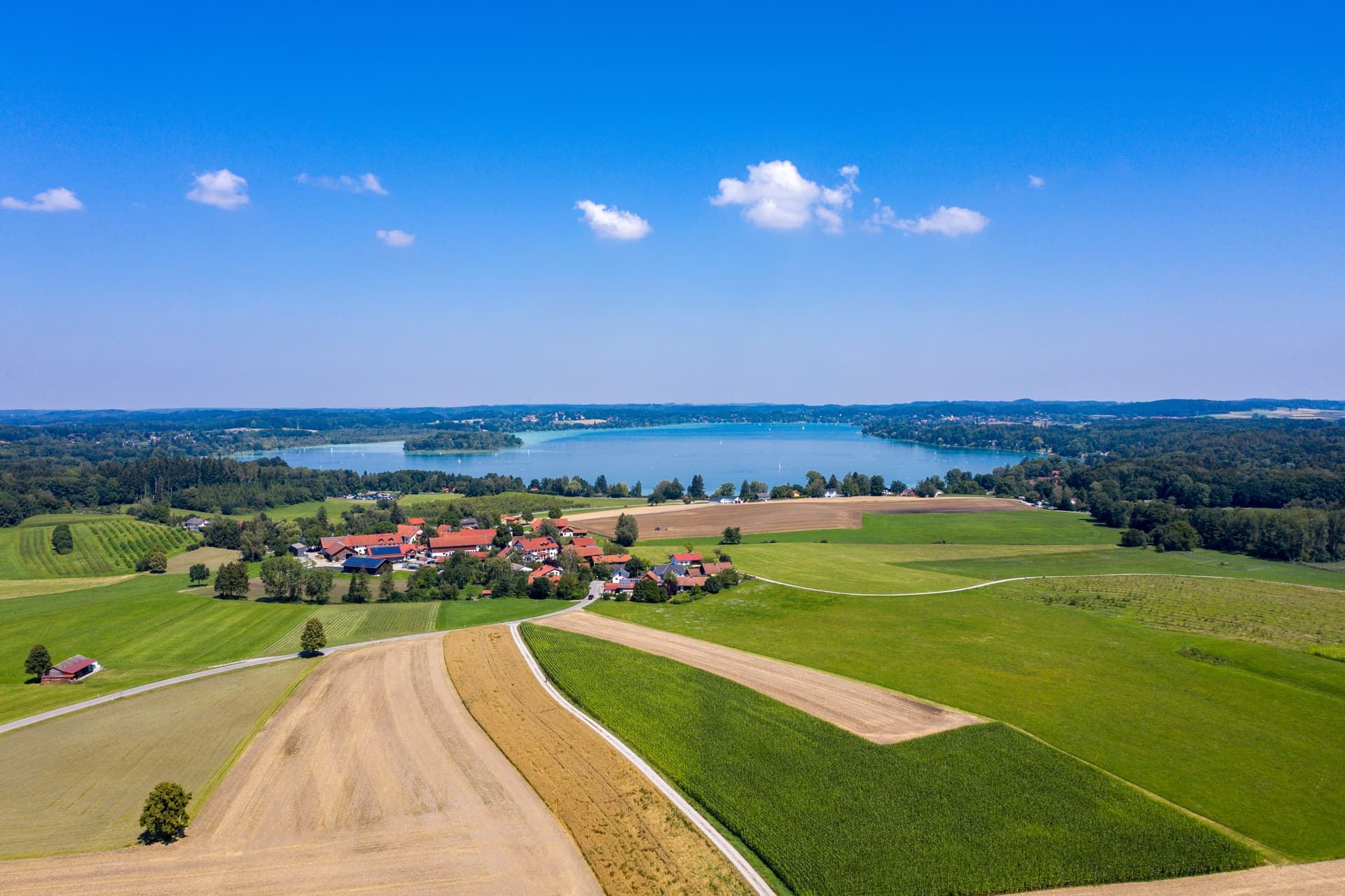 Aerial view of Woerthsee lake with village and patchwork fields under a bright blue sky.