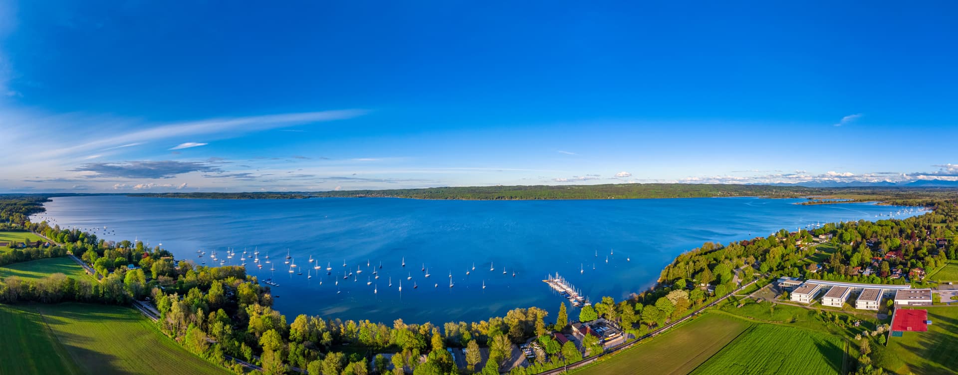 Sailboats moored on Ammersee lake with green shoreline and distant Alps under a bright blue sky.