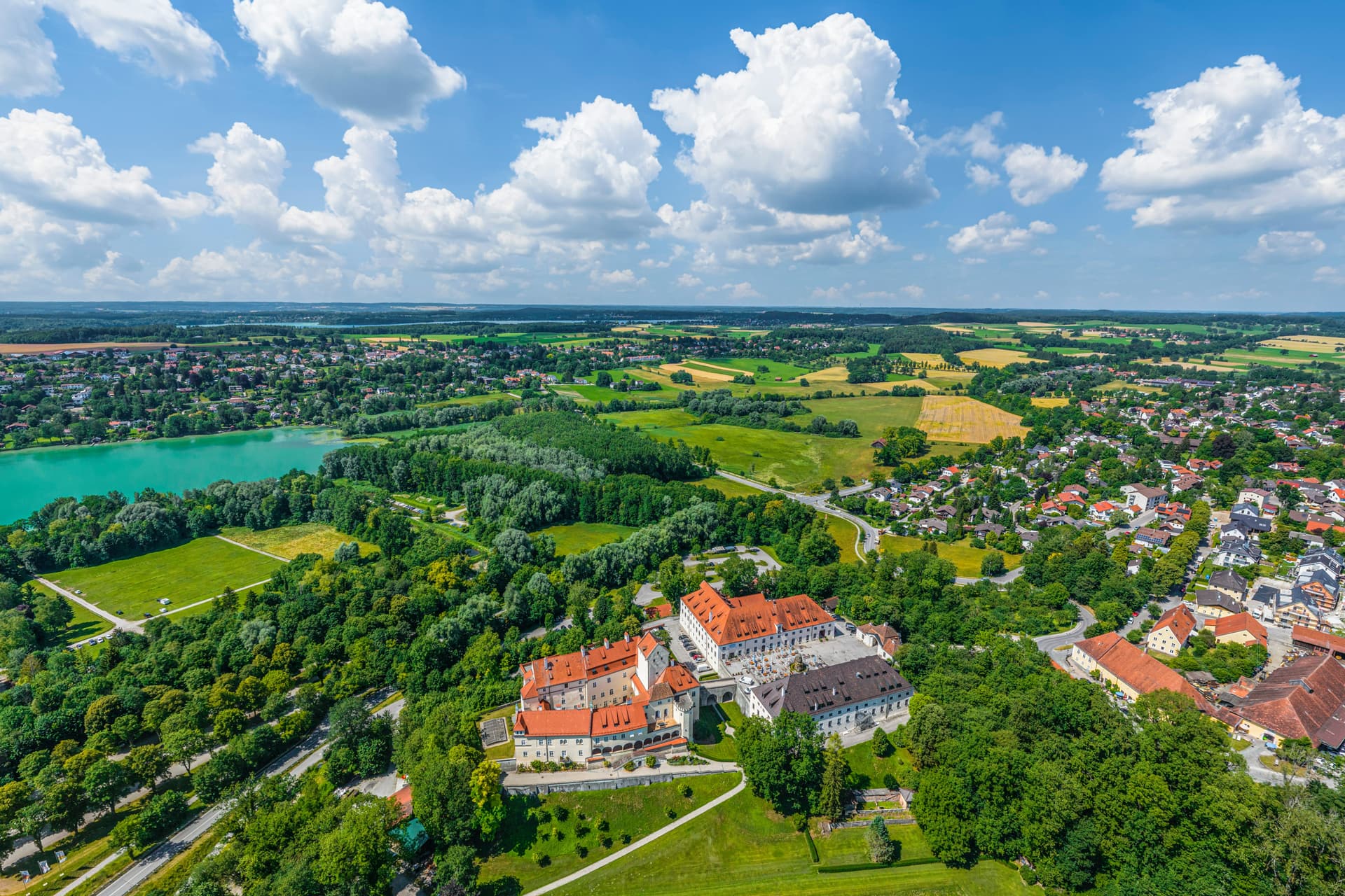 Aerial view of Schloss Pilsensee estate, turquoise lake, and surrounding green landscape under blue sky.