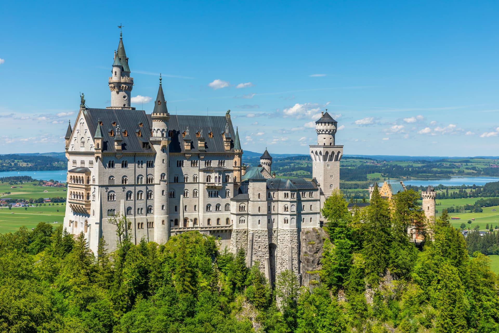 Neuschwanstein Castle on a forested hill overlooking green fields and a lake under a blue sky.