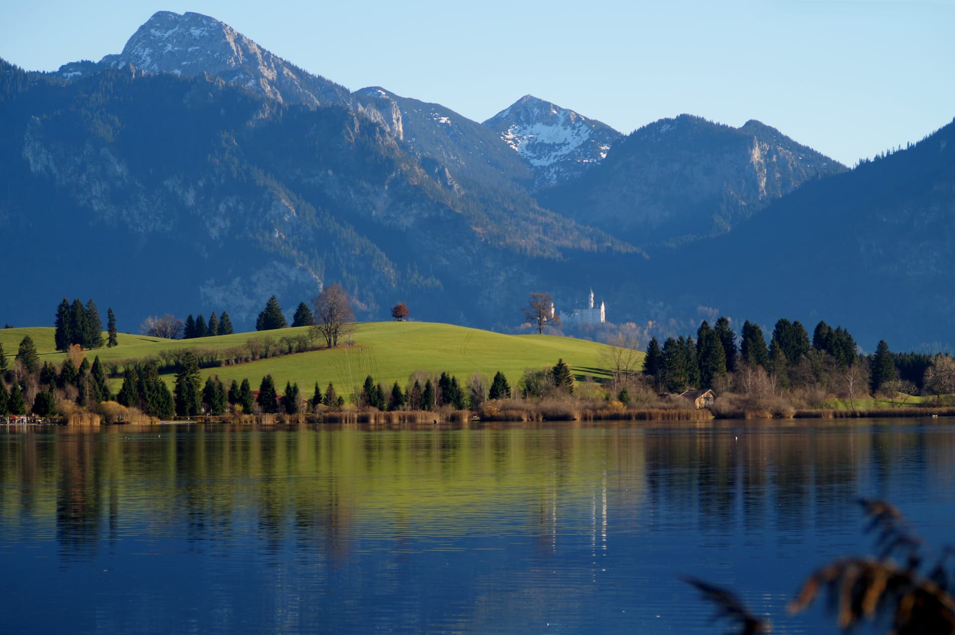 Lake Bannwaldsee with green hills, distant castle, and snow-capped mountains reflected in blue water.