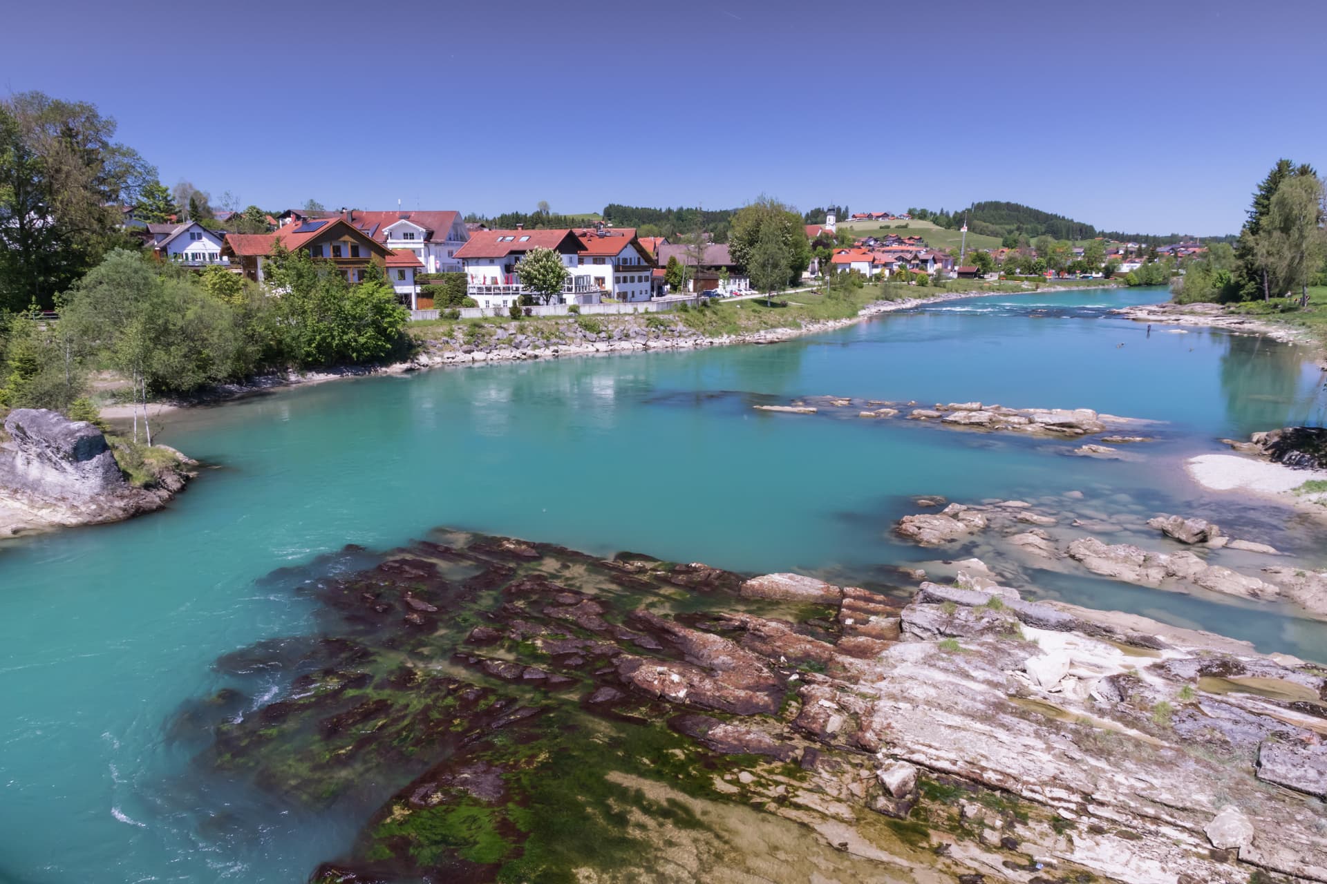 Turquoise river flowing past rocky shallows with a small town nestled against green hills under a clear blue sky.