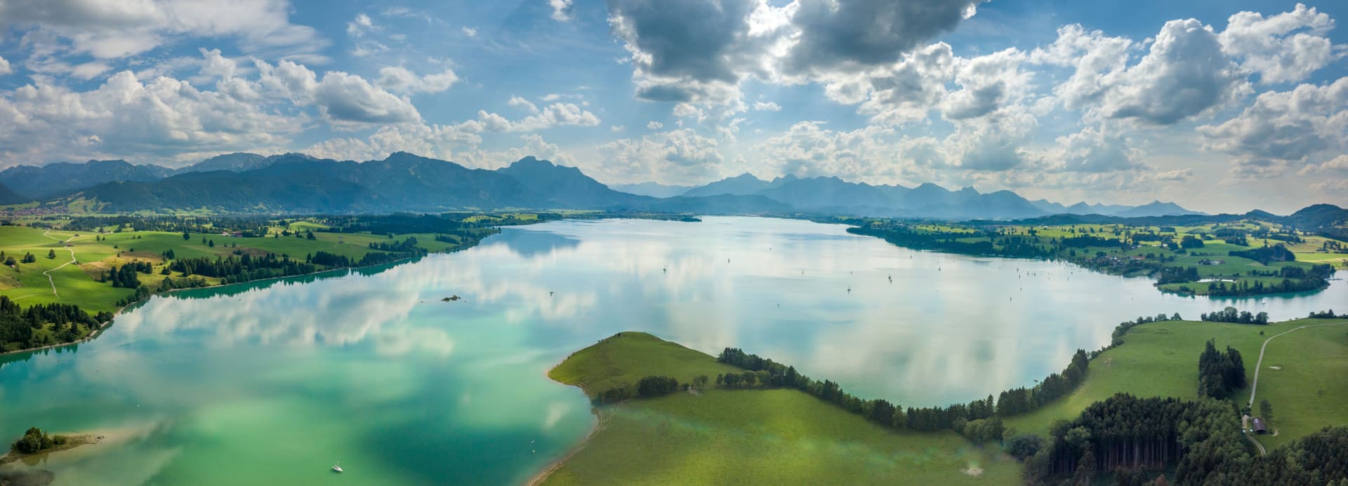 Aerial view of Forggensee lake with turquoise water, green shores, and distant mountains under a cloudy blue sky.