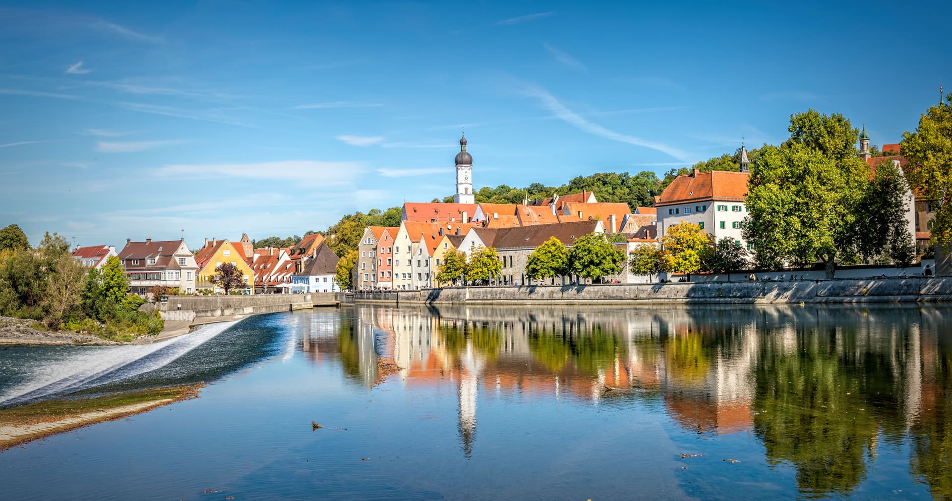 Colorful historic buildings reflected in the calm water of the Landsberg waterfront under a blue sky.