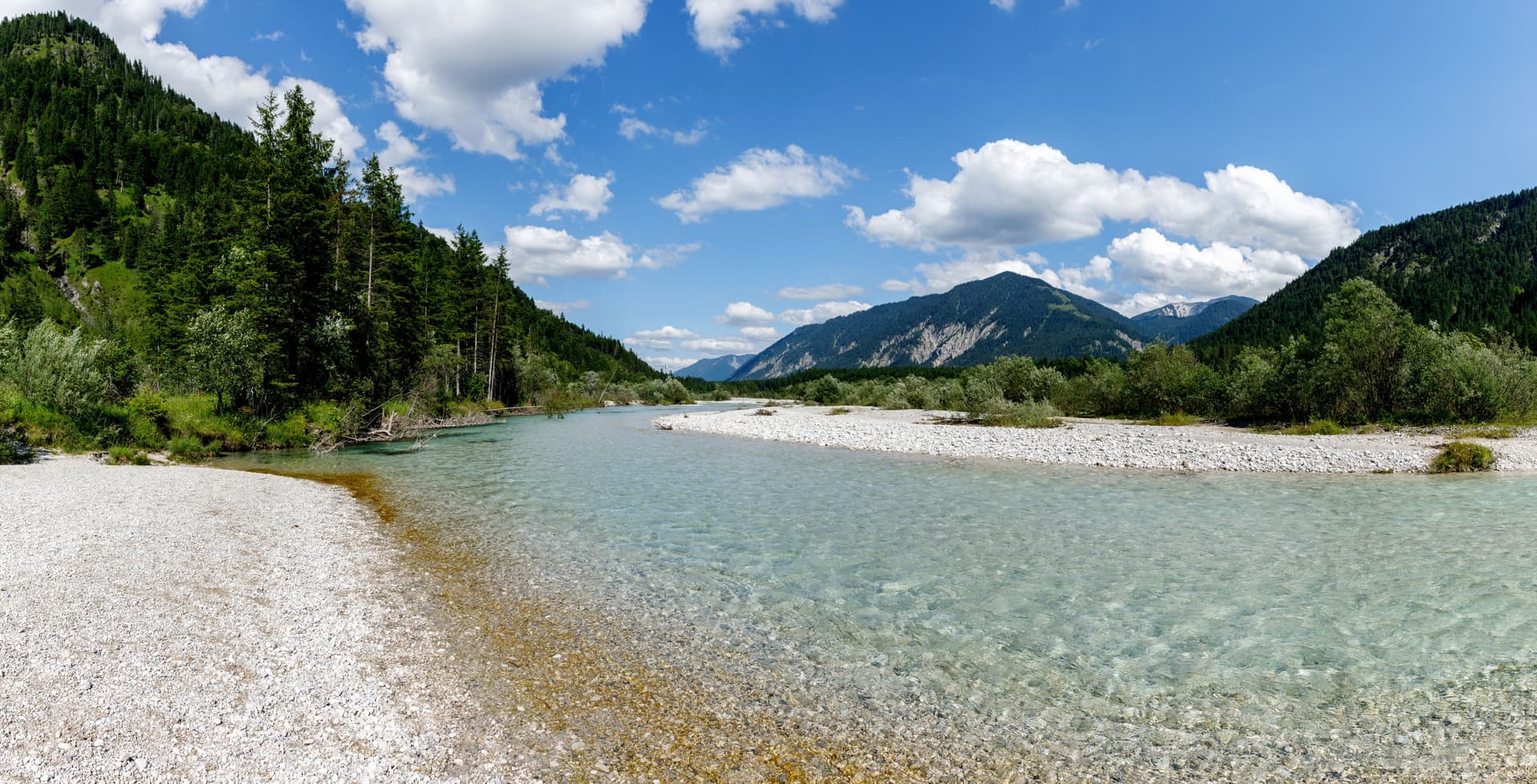 Clear river water flows past a white pebble beach surrounded by forested mountains under a blue sky.
