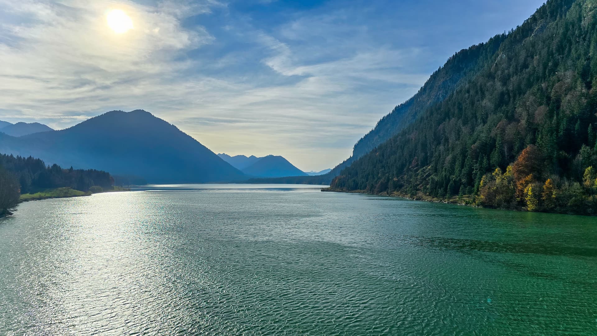 Lake Sylvestersteinsee with sun reflecting on green water, surrounded by forested mountains.