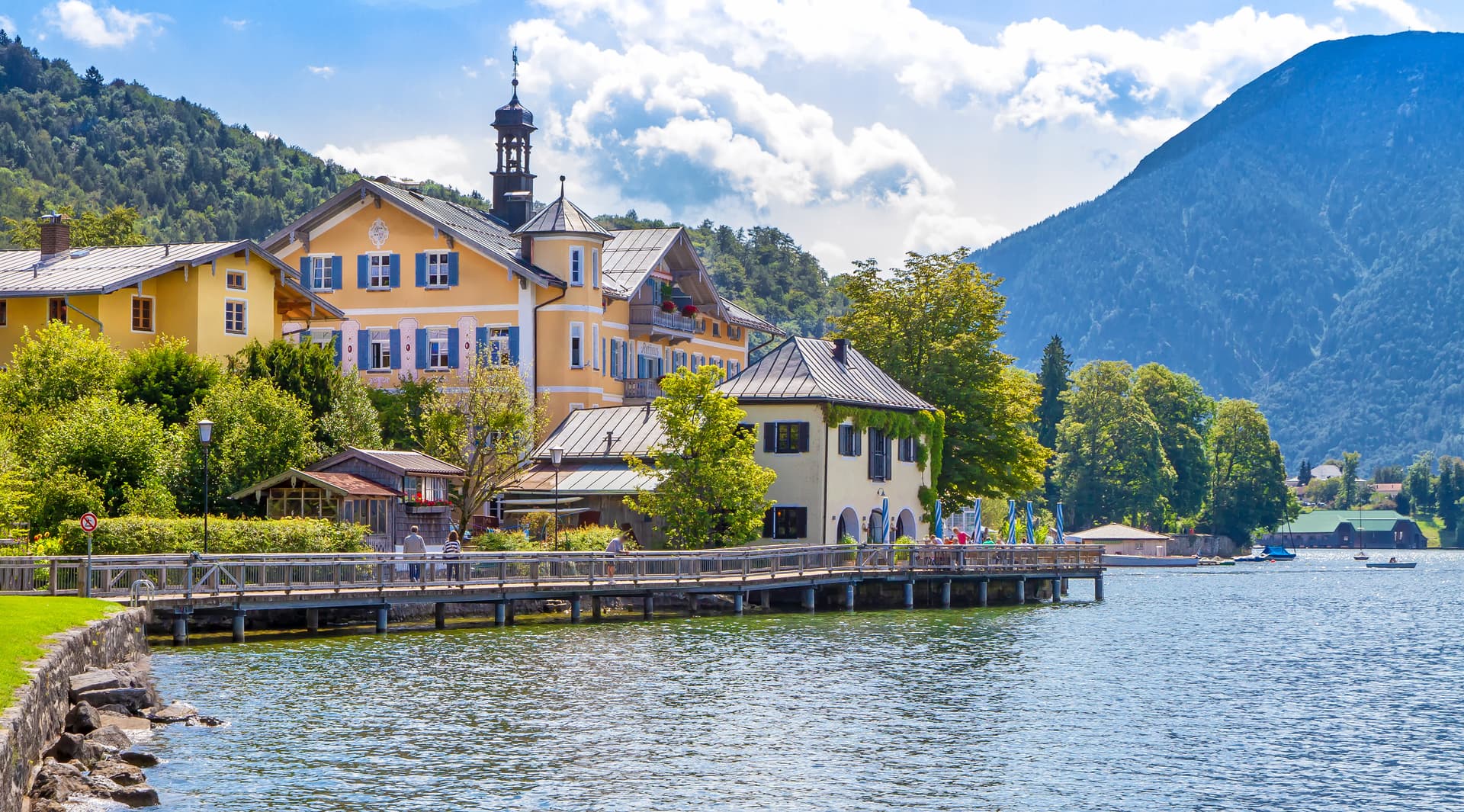 Yellow buildings on Tegernsee lake shore with wooden boardwalk and forested mountains.