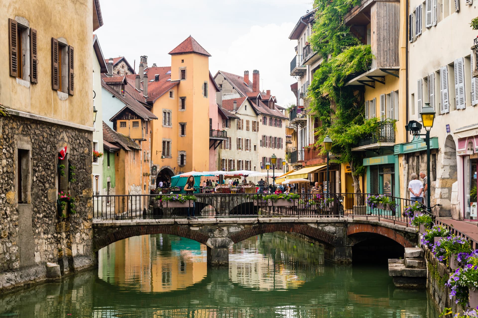 Stone bridge over canal with colorful buildings and outdoor cafe seating in Annecy, France.
